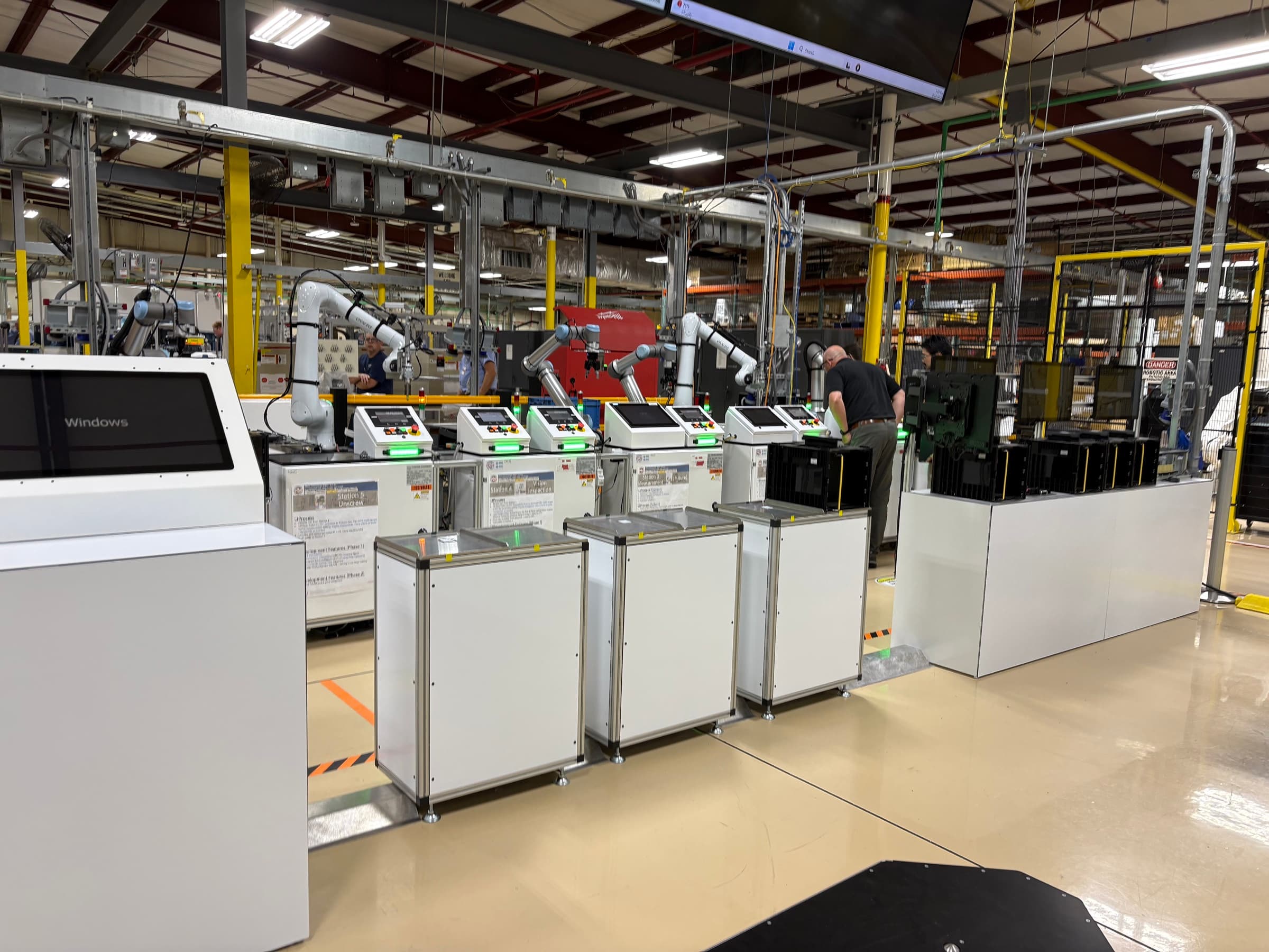 An assembly cell on a bright plant floor: collaborative robot arms, touchscreen terminals at each station, and part carts staged along a marked walkway.